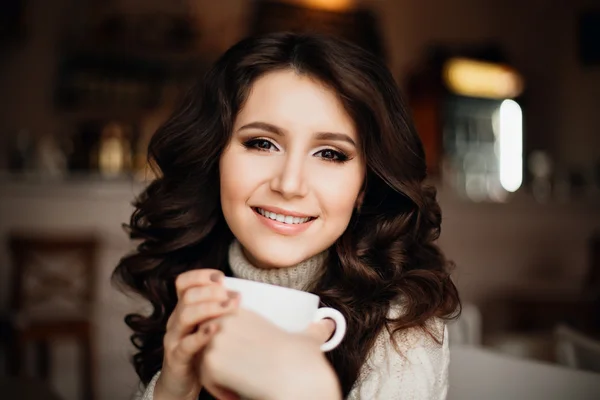 Female model with long hair. Amazing sensual girl with gorgeous make-up is enjoying a delicious coffee or tea, playfully smiling at the camera. Long dark wavy hair, white smile.