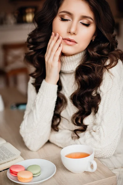 Photos of beautiful brunette with long hair and perfect makeup to wear knitted dress, eyes closed, touches the hand of his face. On the table a Cup of tea or coffee and sweets, macaroon.