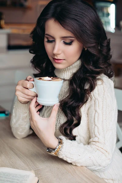 Brunette in a cafe drinking tea, eating sweets, reading a book, looking at the cup, beautiful eyes and gorgeous makeup, wavy hair. Resting after a day of enjoying life. Happy girl. Soft warm colors.