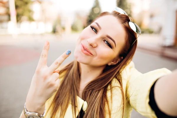 Young sexy woman with bright make-up formic blond hair makes selfie sending a kiss, hipster street portrait. Shows a peace sign, taking pictures of herself.