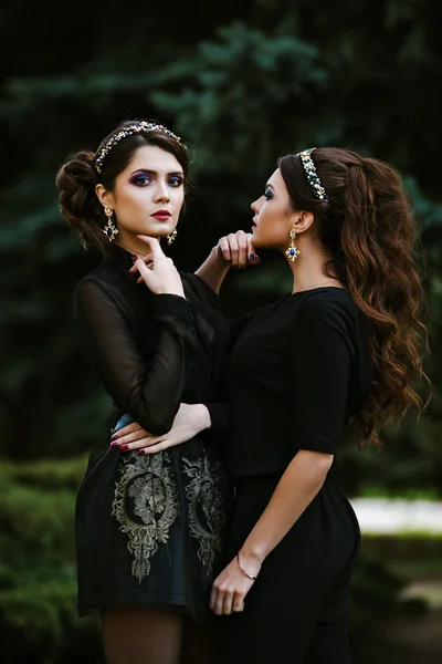 Two young beautiful fashionable woman in black clothes posing for the photographer. Headbands on the head, in the ears of stylish earrings. Expensive jewelry. curly hairstyles.