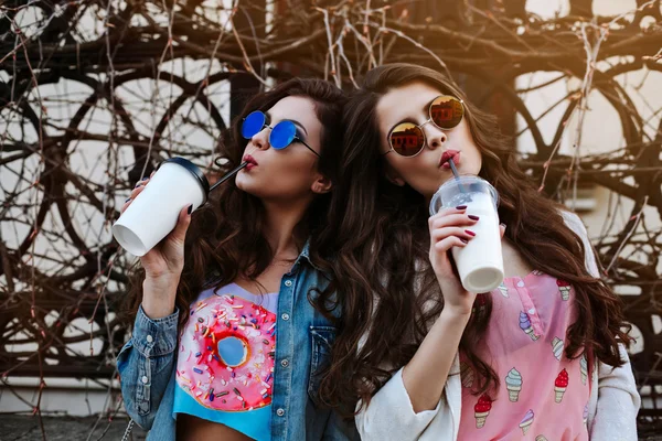 Outdoor fashion lifestyle portrait of two young beautiful women, dressed in denim outfit, mirrored sunglasses, enjoy a stroll, drink coffee, bright stylish clothes, accessories