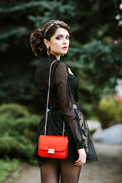 Lifestyle portrait of young stylish woman goes in the city with a red trendy bag. On the girl shoulder clutch bag, wearing a black dress, wearing a headband with precious stones. Earrings, jewelry.