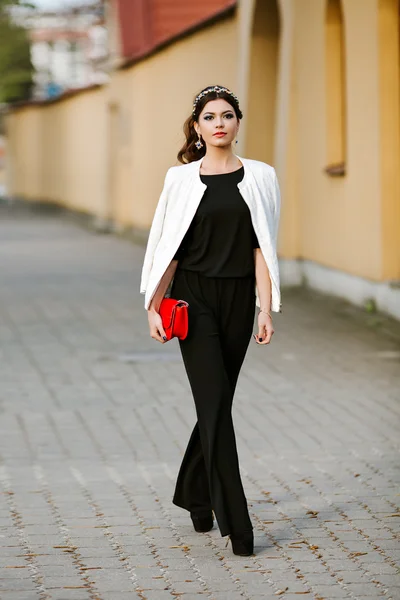 Young stylish woman goes in the city with a red trendy bag. On the shoulder of the girl bag, dressed in a black Trouser suit, white jacket, bezel gemstones. Earrings, jewelry.