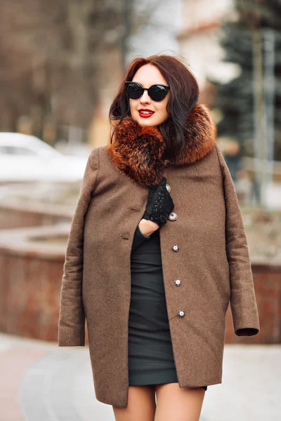 Beautiful sexy glamorous brunette girl in an elegant green dress and fur coat with a fluffy fur, gloves and stylish sunglasses outdoors. young woman with chic long dark hair and trendy makeup