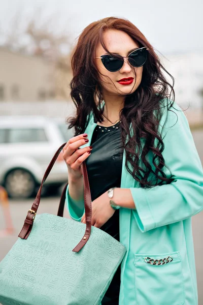 Amazing beautiful girl with dark hair. Fashion handbag, long coat mint color. Stylish sunglasses. Red plump lips, beautiful makeup.