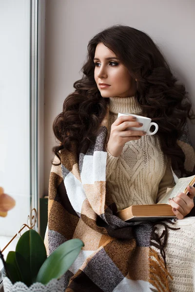 Beautiful young woman drinking tea and reading a book, covered with a warm blanket,dressed in a white knit dress at the window.