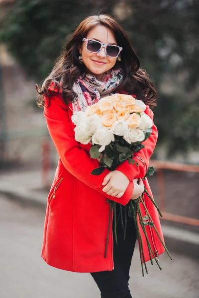 Happy young woman in bright coat with flowers goes through the city. Beautiful girl holding rose bouquet and smiling. A woman with dark hair wearing sunglasses.
