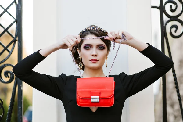 Lifestyle portrait of young stylish woman goes in the city with a red trendy bag. On the girl shoulder clutch bag, wearing a black dress, wearing a headband with precious stones. Earrings, jewelry.