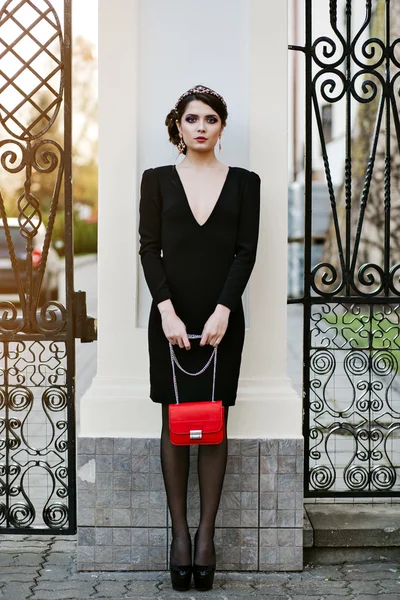 Lifestyle portrait of young stylish woman goes in the city with a red trendy bag. On the girl shoulder clutch bag, wearing a black dress, wearing a headband with precious stones. Earrings, jewelry.
