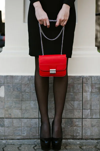 Lifestyle portrait of young stylish woman goes in the city with a red trendy bag. On the girl shoulder clutch bag, wearing a black dress, wearing a headband with precious stones. Earrings, jewelry.