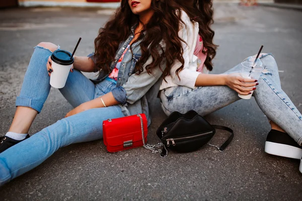 Two fashionable young women sit side by side, dressed in a denim outfit, bright t-shirts, coffee in hand, fashion handbags.