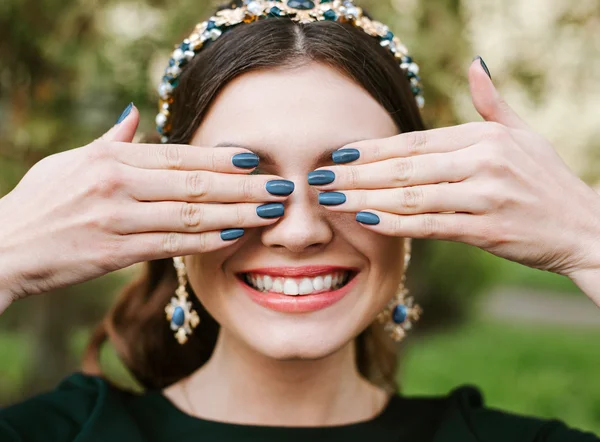 Fashion, beauty, tenderness. Young happy woman with a bright manicure smile wide,white smile,straight white teeth. The girl covers her face with hands. Hair band, earrings, blue nail Polish.