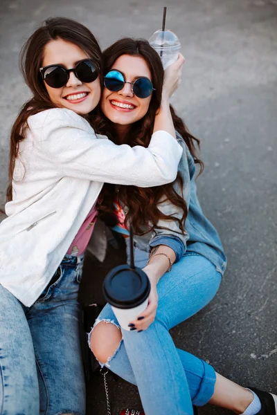 Outdoor fashion lifestyle portrait of two young beautiful women, dressed in denim outfit, mirrored sunglasses, enjoy a stroll, drink coffee, bright stylish clothes, accessories