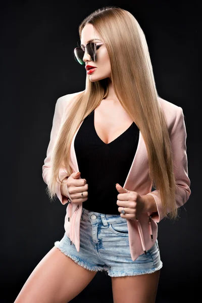 Fashionable stylish woman in denim outfit and a beautiful coat looking to the side at the mirrored sun glasses. The girls red lips. Studio photo on a black background.