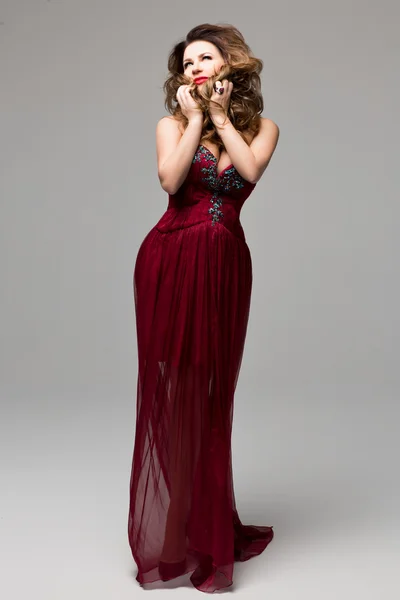 Beautiful young female I in a long red dress with precious stones on a gray background. Clothing catalog, catalog design. Chic design of the dress. Evening makeup, red lips, wavy hair.