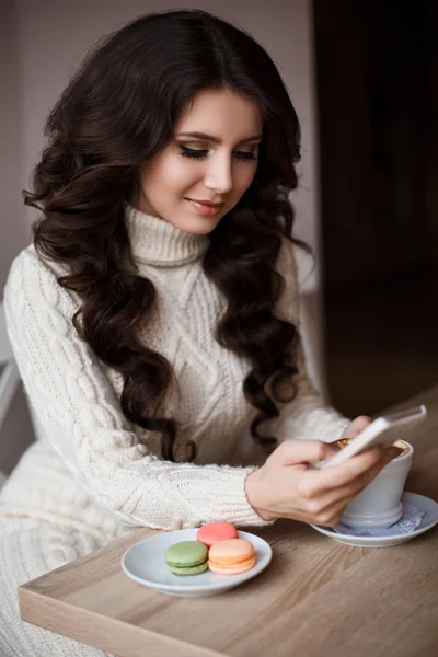 Inside portrait of smiling woman with long hair writes the message. On the table coffee,sweets,and macaroons. Communication, smile, joy, rest.