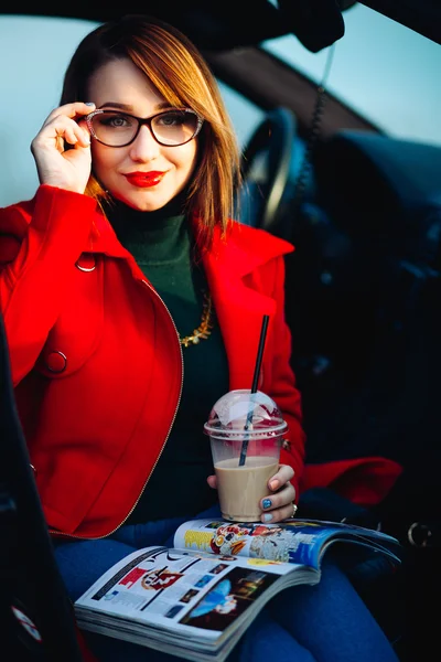 Lifestyle portrait of a remarkable young woman posing with glasses for vision. Girl Drinking coffee and reading a magazine, sitting in the car. Dressed in a bright coat and denim outfit.