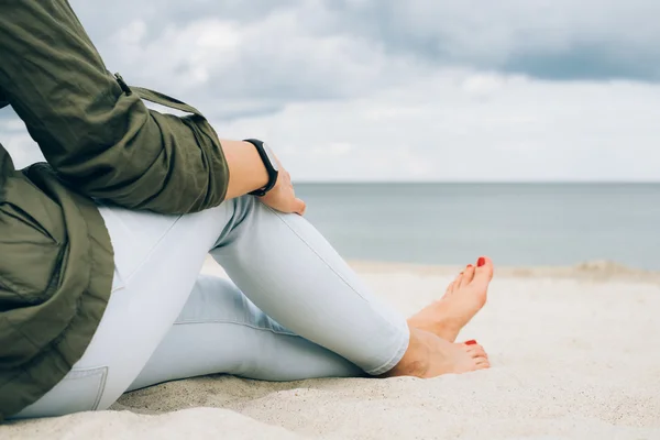 Woman in green jacket and jeans sits on a beach and looks at sea