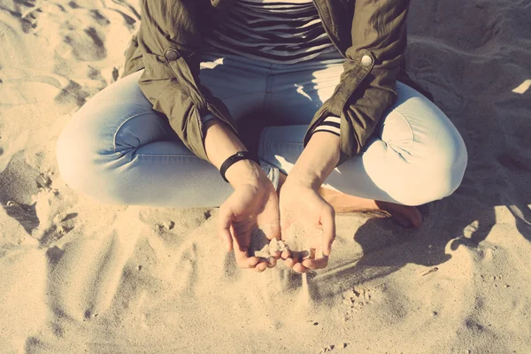 Woman in coat and jeans sitting on the beach cross-legged, in he