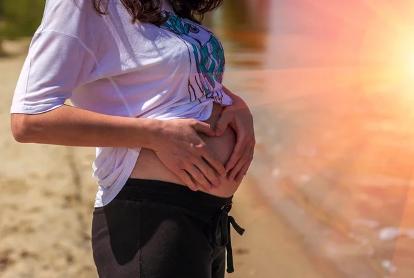 Pregnant woman touches her belly hands making a heart on the bea