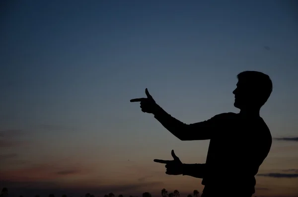 Silhouette against a sunset sky of an adult man with a backpack pointing with his arm straight in front of him.