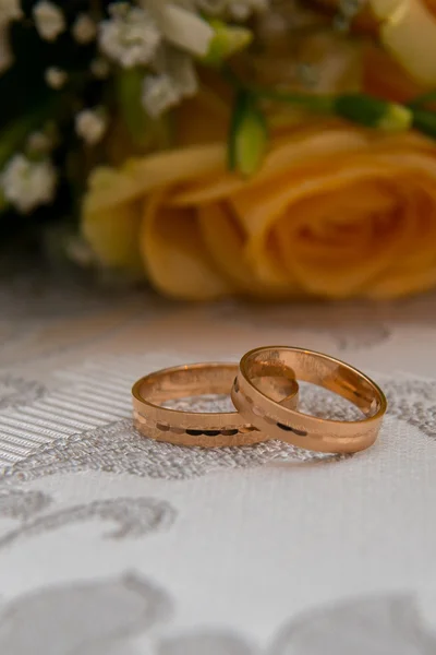 Two gold wedding rings lie on the table near the bouquet of orange roses and white flowers.
