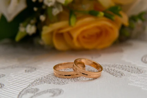 Two gold wedding rings lie on the table near the bouquet of orange roses and white flowers.