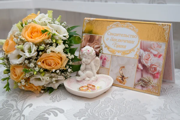 Two gold wedding rings lie on a platter in a rose shape with the angel sculpture near the bride\'s bouquet of orange roses and white flowers. The certificate of marriage.