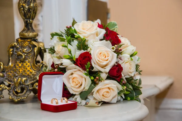 Two wedding gold rings with diamonds are in the red box near the bride\'s bouquet of red and white roses.