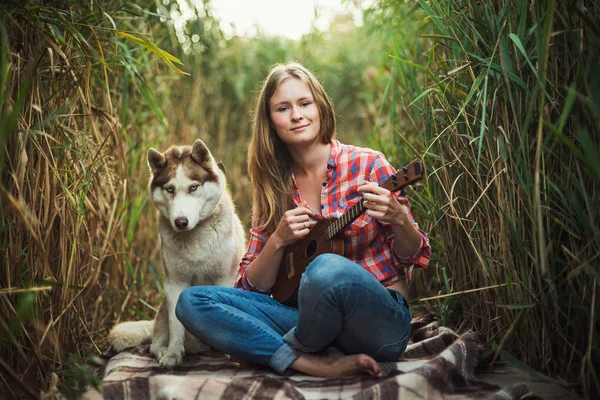 Young caucasian woman playing ukulele. Female with siberian husky dog playing guitar outdoors