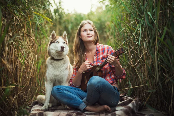 Young caucasian woman playing ukulele. Female with siberian husky dog playing guitar outdoors