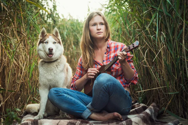 Young caucasian woman playing ukulele. Female with siberian husky dog playing guitar outdoors