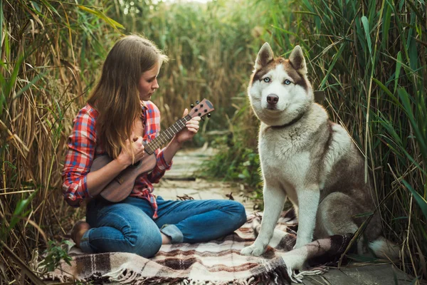 Young caucasian woman playing ukulele. Female with siberian husky dog playing guitar outdoors