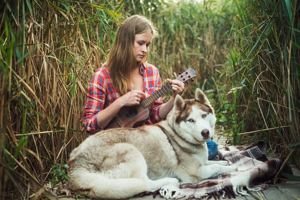 Young caucasian woman playing ukulele. Female with siberian husky dog playing guitar outdoors