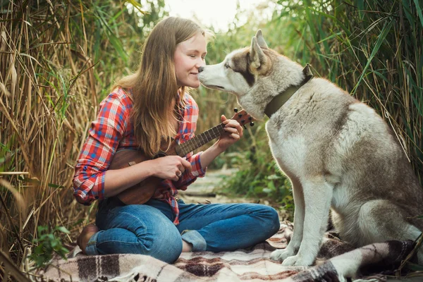 Young caucasian woman playing ukulele. Female with siberian husky dog playing guitar outdoors