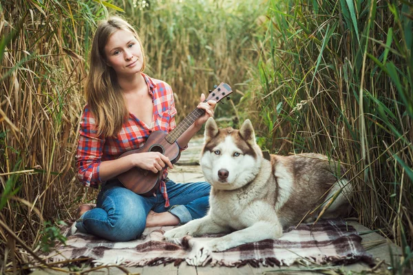 Young caucasian woman playing ukulele. Female with siberian husky dog playing guitar outdoors