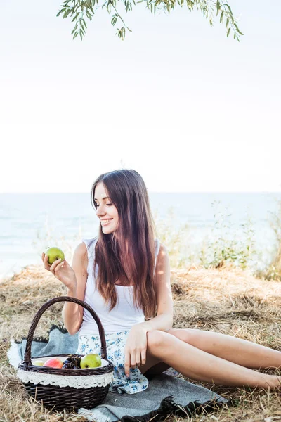 Young caucasian female on seaside with basket with fruits. Girl eating fruits, healthy eating and healthy living concept