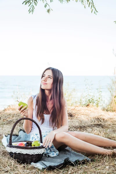 Young caucasian female on seaside with basket with fruits. Girl eating fruits, healthy eating and healthy living concept