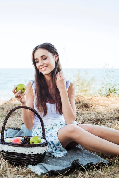 Young caucasian female on seaside with basket with fruits. Girl eating fruits, healthy eating and healthy living concept