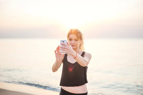 Young beautiful caucasian fitness model taking pictures of herself with mobile phone camera on beach