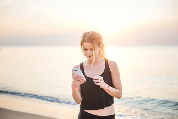 Young beautiful caucasian fitness model taking pictures of herself with mobile phone camera on beach