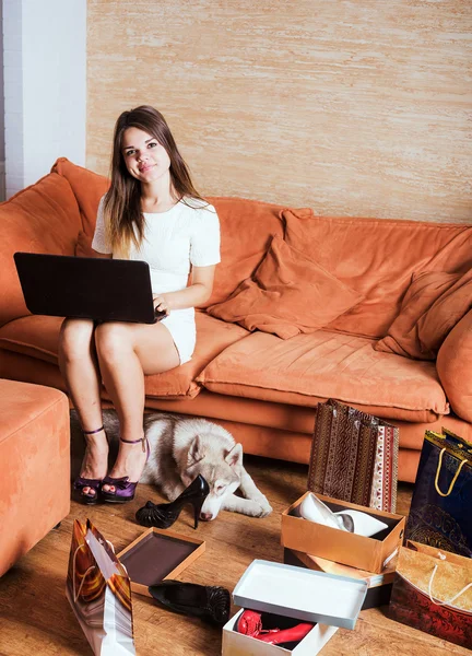 Young caucasian female with laptop and shopping bags in living room. Woman tired of shopping. Female and little husky dog after shopping at home