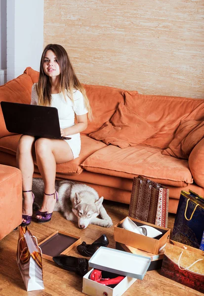 Young caucasian female with laptop and shopping bags in living room. Woman tired of shopping. Female and little husky dog after shopping at home