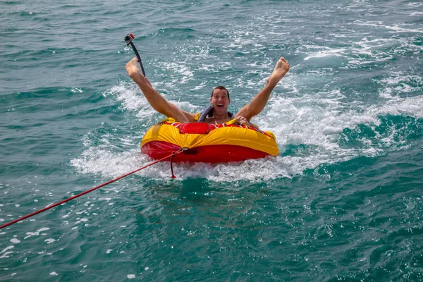 Man sitting in inflatable ring towed by a boat in the water and making faces to the camera and holding Go Pro camera
