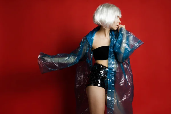 The photo in landscape orientation. Beautiful girl in a white wig, black underwear and a raincoat in the studio on a red background. Sexy young woman holding her hand near head and looking at the camera.