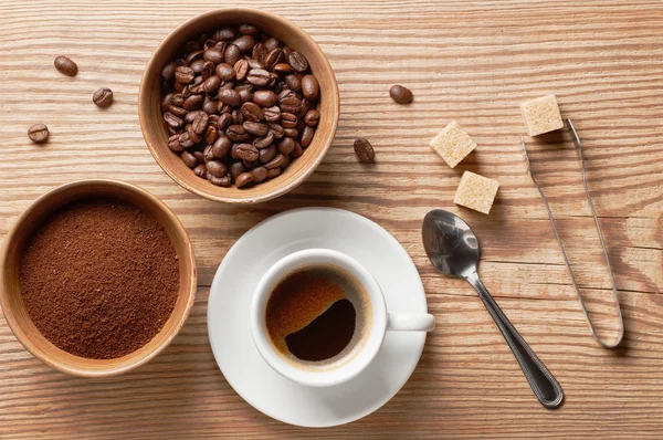 Coffee beans, ground coffee and cup of coffee on wooden table with spoon, sugar tongs, cane sugar cubes and coffee beans, top view