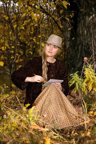 Beautiful young blonde woman sitting outdoors in last sunbeams at autumn sunset paints a sketch in notepad