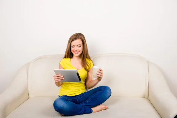 Young happy woman with a cup holding tablet sitting on the sofa