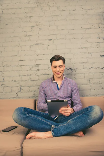 Handsome young man sitting on couch and reading news on tablet